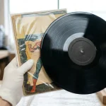 Colecționar purtând mănuși albe de bumbac, ținând cu grijă un disc de vinil și o revistă de benzi desenate în stare perfectă, ilustrând conceptul de 'Mint Condition